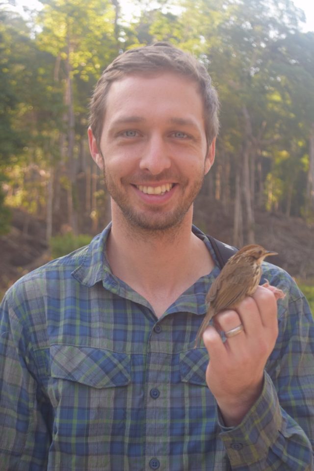 Ecologist Grant Connette smiles in a forest while gently holding a small brown bird.