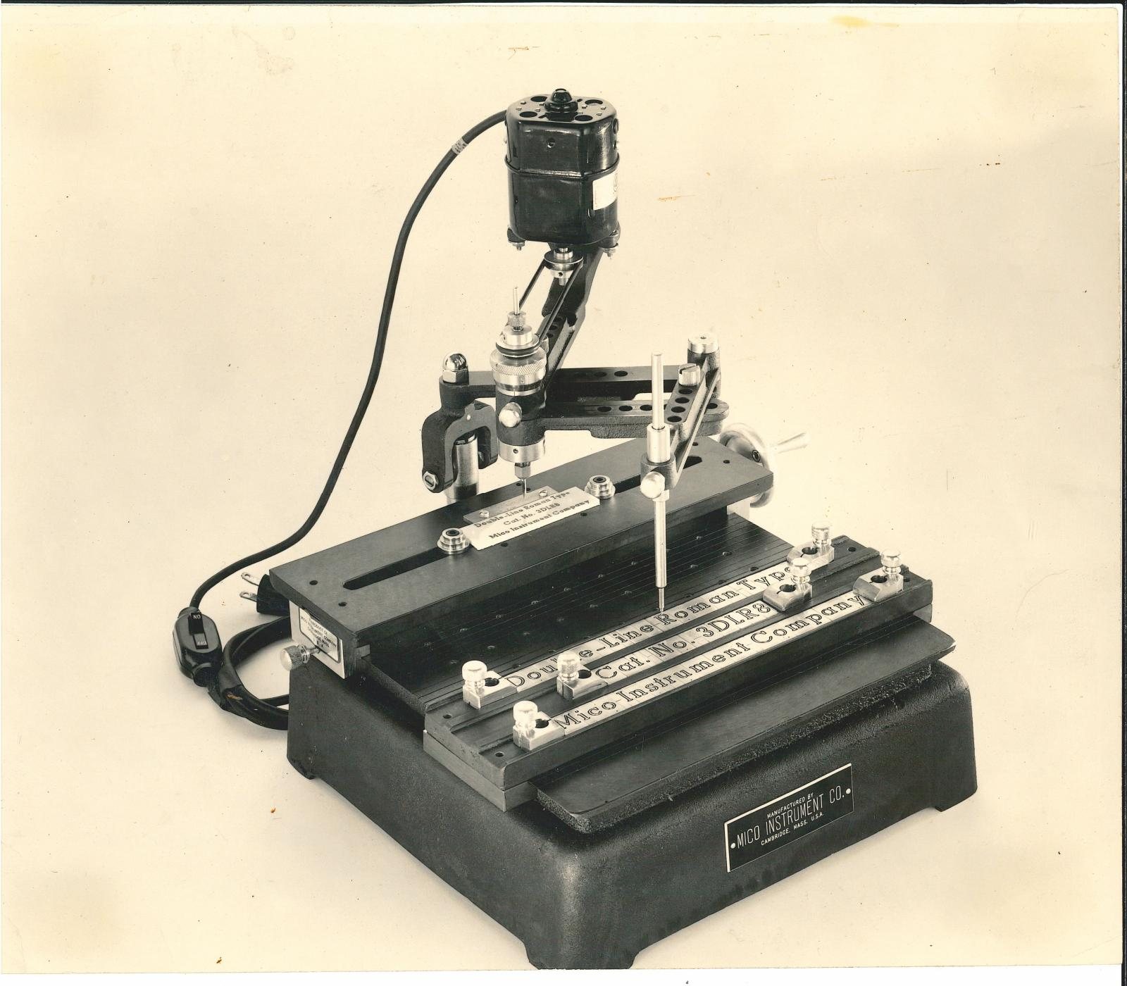 The Mico Standard Engraver: a cast-iron bench-top pantograph machine with an electric motor mounted above the arm and engraved nameplates held in the copy carrier