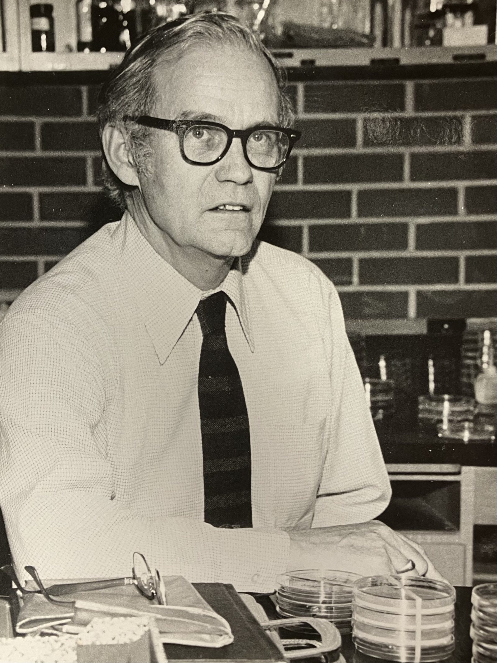 Black-and-white photograph of Dr. Lindsay S. Olive in glasses and a tie, seated at a lab bench with stacked petri dishes
