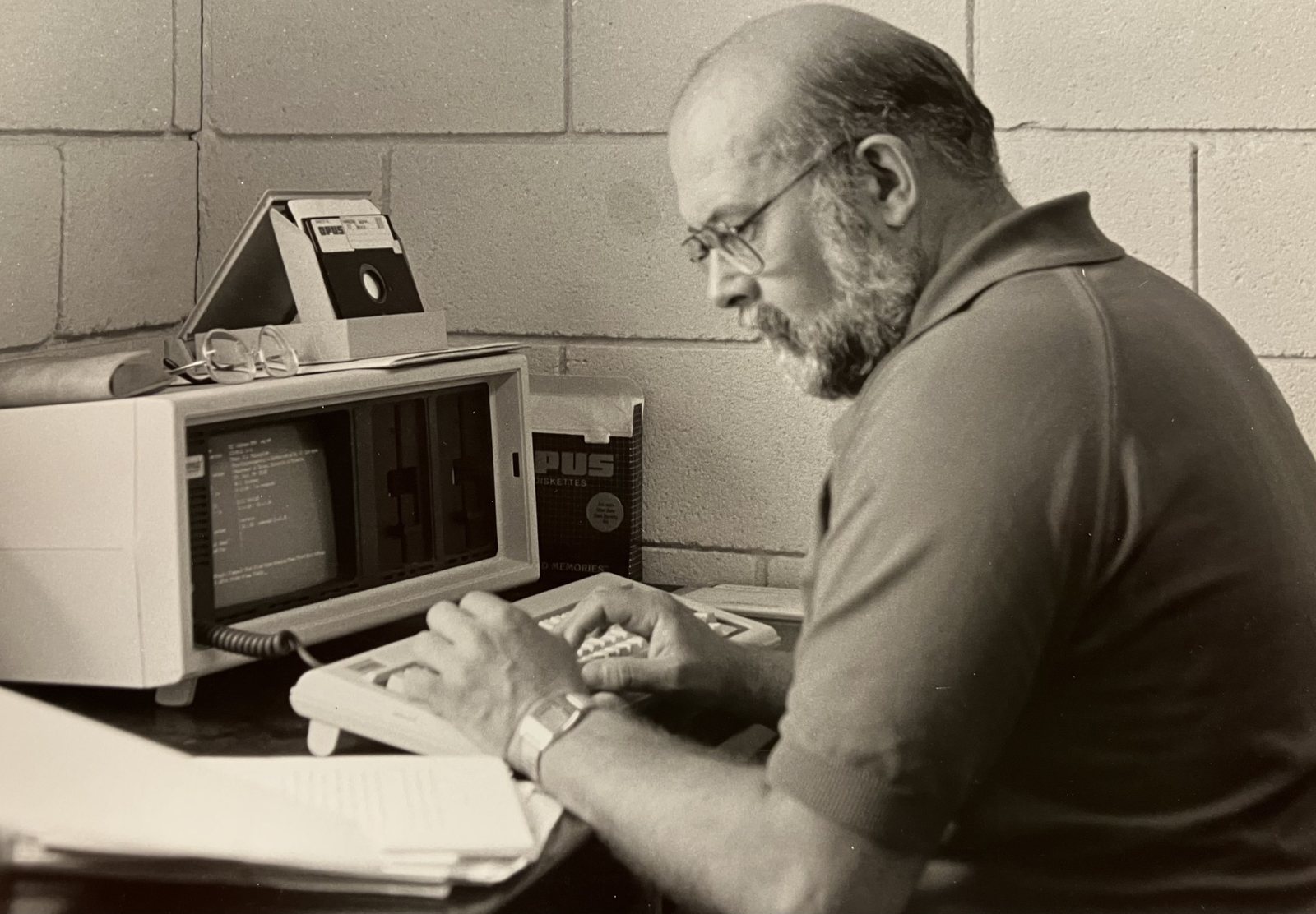Black-and-white photograph of Dr. Ronald H. Petersen, bearded and in glasses, typing at an early desktop computer with floppy disks beside it