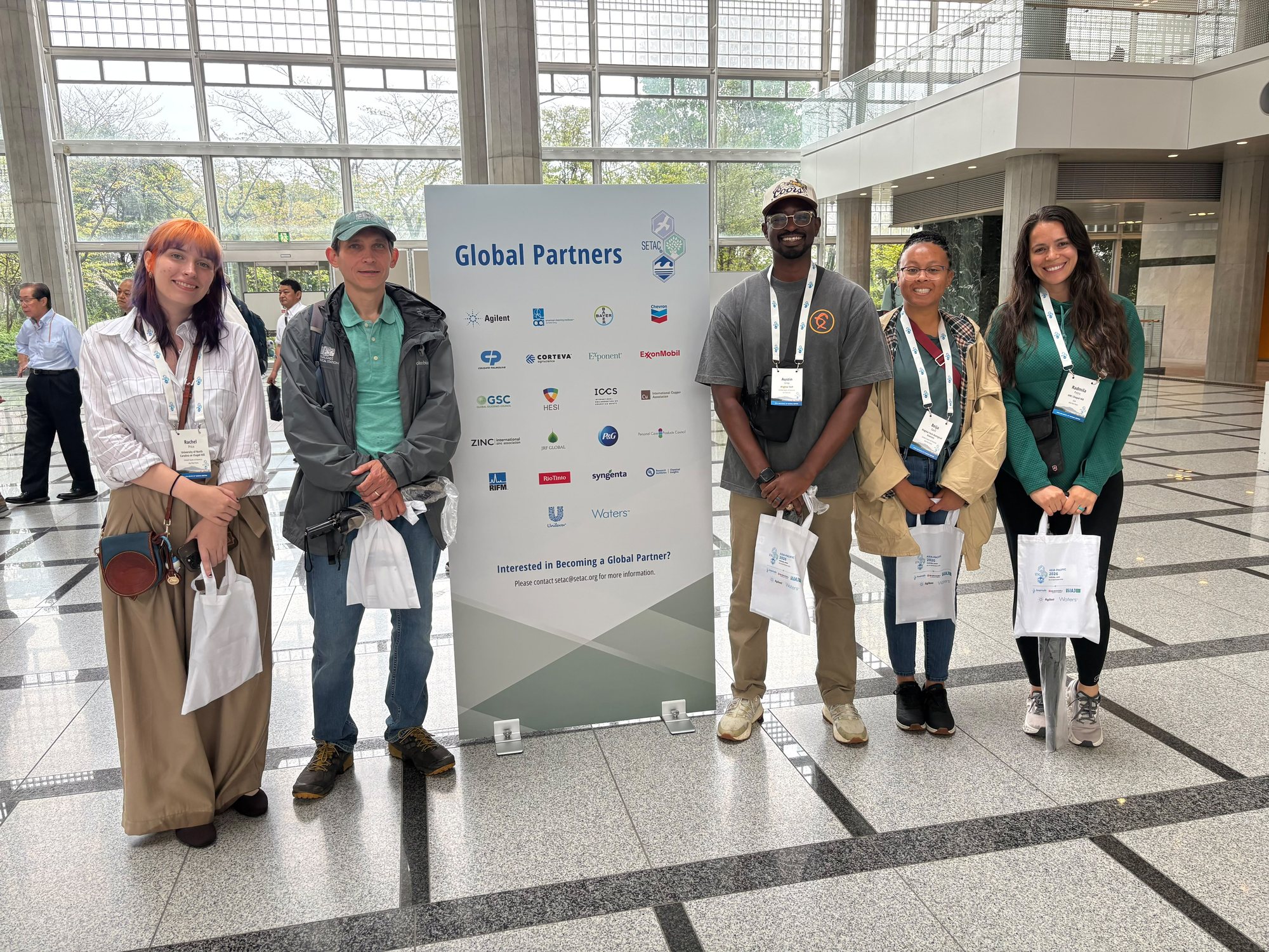 Rachel Price, Jason Love, Austin Gray, Beija Gore and Rada Petric stand together, smiling, in front of a Global Partners banner in the bright conference hall in Tsukuba, Japan, conference badges and tote bags in hand.
